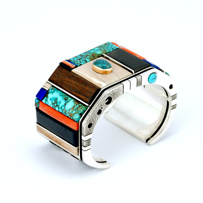 Navajo jeweler Wes Willie created a sterling silver inlay bracelet featuring turquoise, coral, lapis, ironwood, fossilized ivory, jet, and fine-line silver bars. The bracelet includes a Lone Mountain turquoise stone set in gold and a hallmark inside.