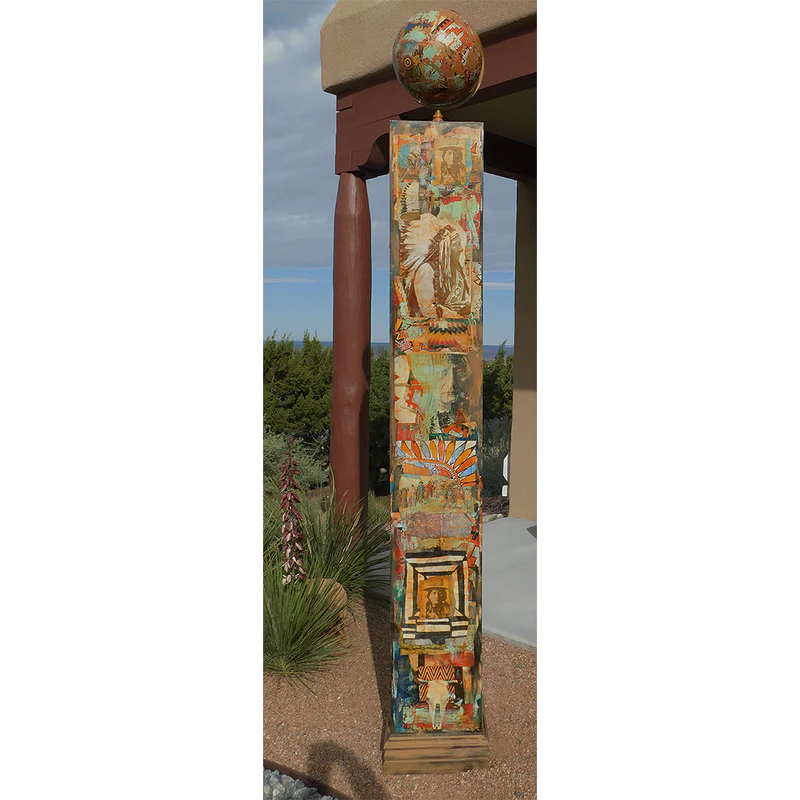101x14 Native Totem by Dave Newman is a tall mixed-media totem sculpture featuring layered imagery, geometric patterns, and earthy Southwestern colors. The vertical piece includes vintage-style portraits, symbolic motifs, and textured collage elements in turquoise, rust, gold, cream, and black, topped with a decorated sphere. Installed outdoors near desert landscaping and adobe-style architecture, the work has a ceremonial, architectural presence.