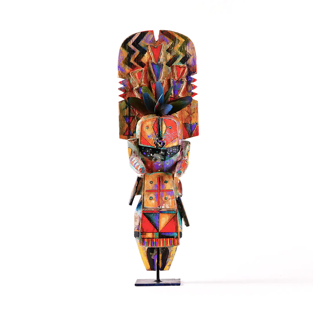 Colorful carved wooden figure on a stand, decorated with bold geometric patterns, zigzags, triangular motifs, and feather-like elements, with a stylized human face and body in bright red, orange, blue, purple, and black against a white background.