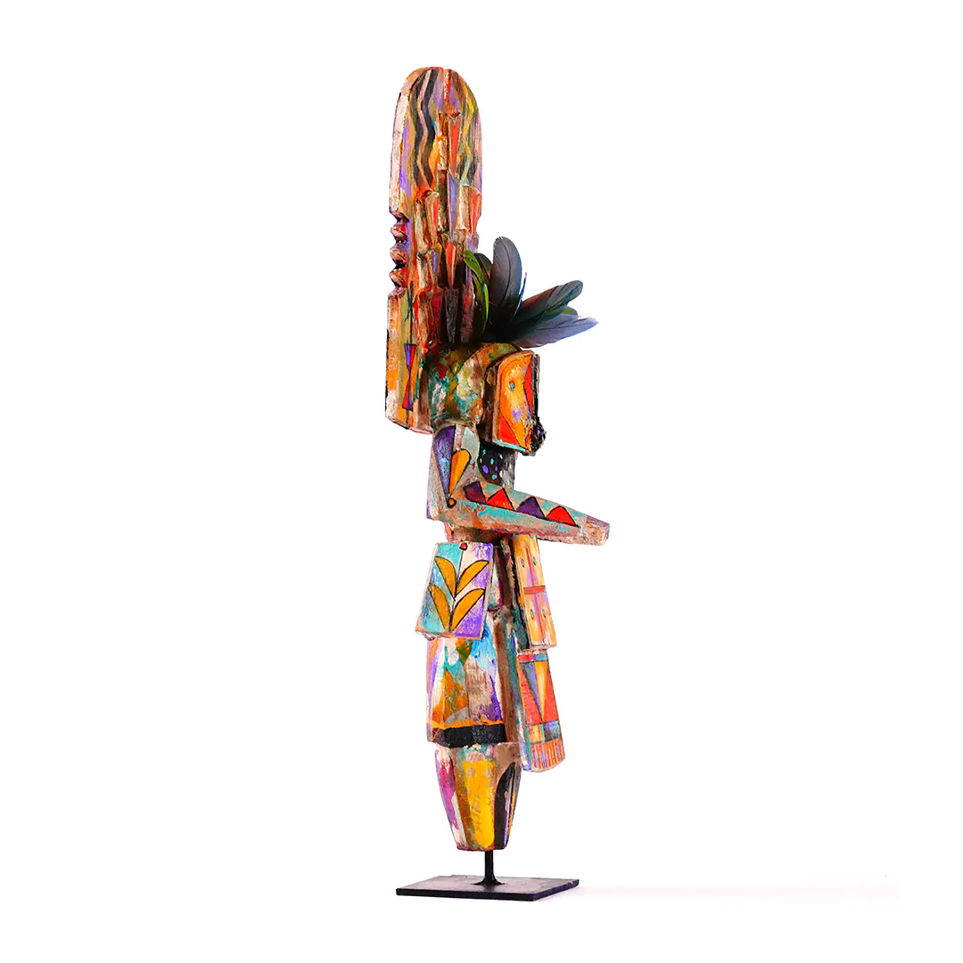 Colorful Navajo Yei sculpture shown in side view, hand-carved in wood with geometric painted details, feather accents, and a tall stylized figure on a stand.