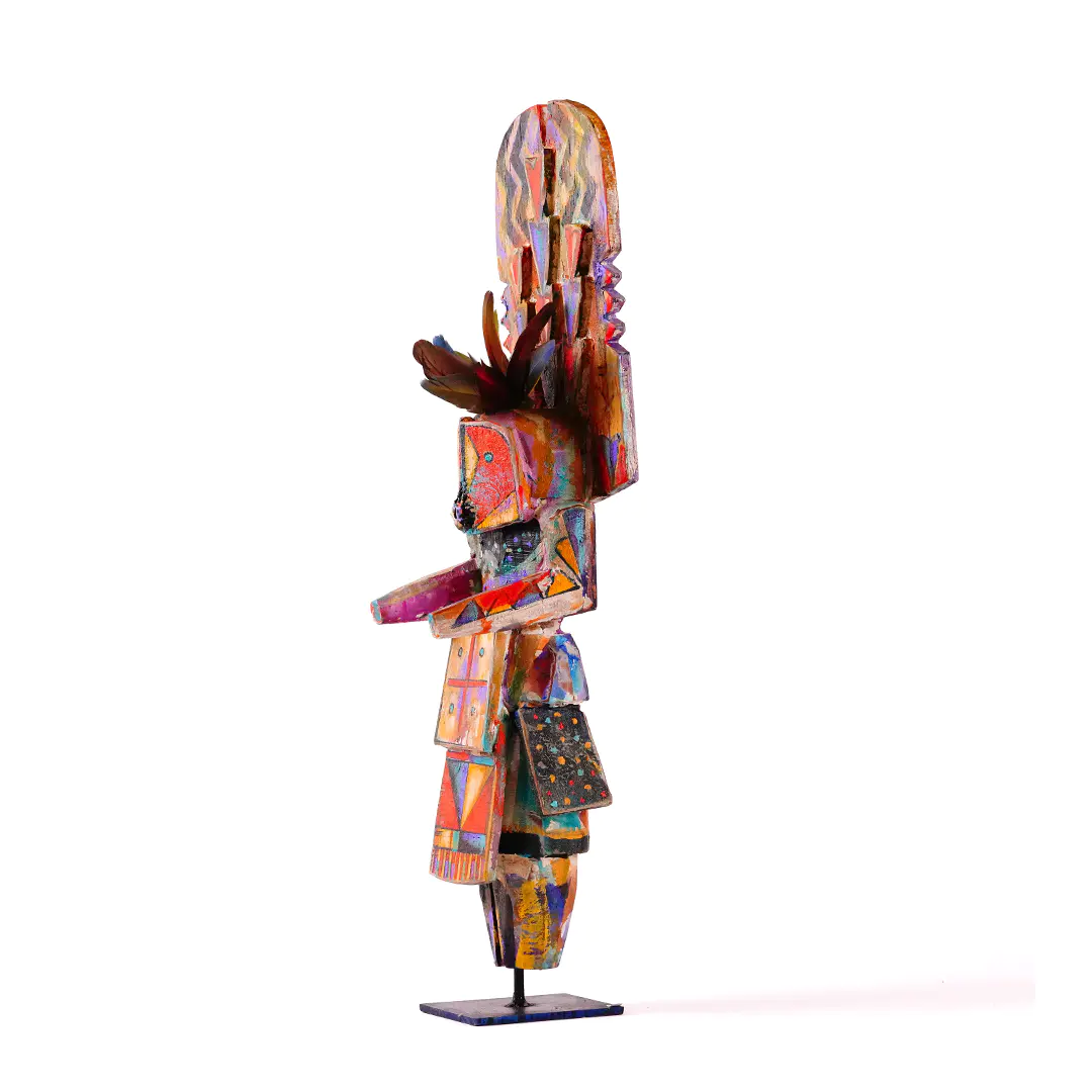 Colorful Navajo Yei sculpture shown in profile, hand-carved in wood with painted geometric details, feather accents, and a stylized figure displayed on a stand.