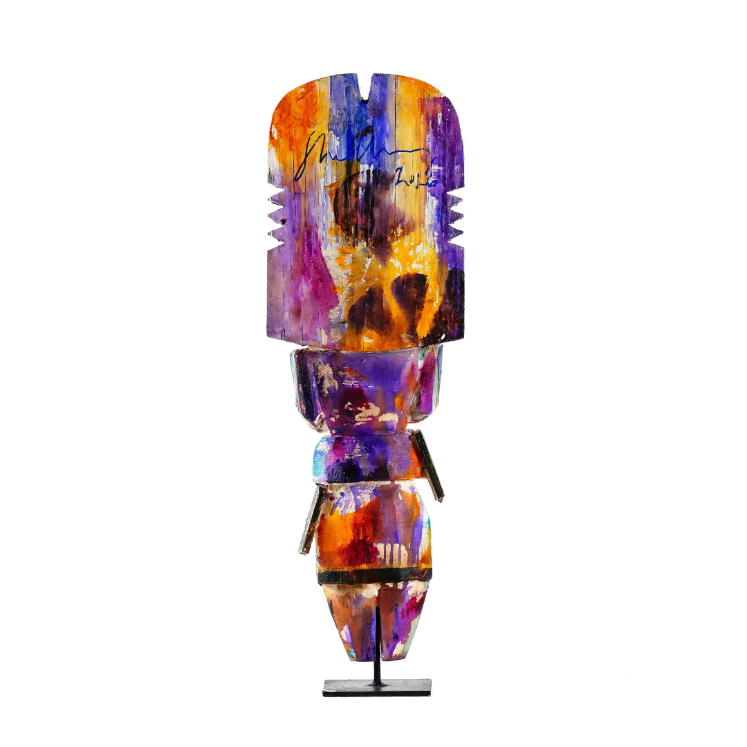 Colorful carved wood figure sculpture, circa 2026, shown from the back with abstract purple, orange, blue, and gold painted details on a stand against a white background.