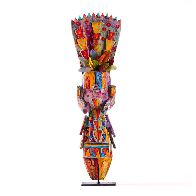 Colorful carved wood figure sculpture with geometric painted patterns, feather accents, and a stylized human form displayed upright on a stand against a white background.