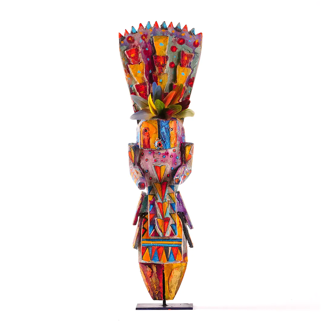 Colorful carved wood figure sculpture with geometric painted patterns, feather accents, and a stylized human form displayed upright on a stand against a white background.