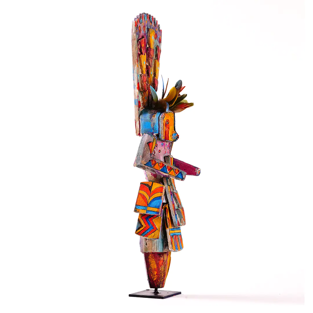 ide view of a colorful stylized Navajo Yei wood sculpture by Sheldon Harvey, featuring geometric hand-painted patterns, a tall crest, and parrot feather accents on a metal stand.