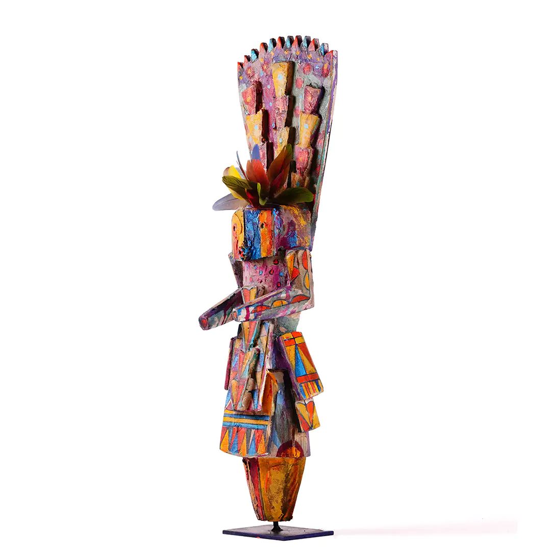 Side view of a brightly painted stylized Navajo Yei wood sculpture by Sheldon Harvey with geometric patterns, a tall headdress, and multicolored parrot feathers, mounted on a metal stand.