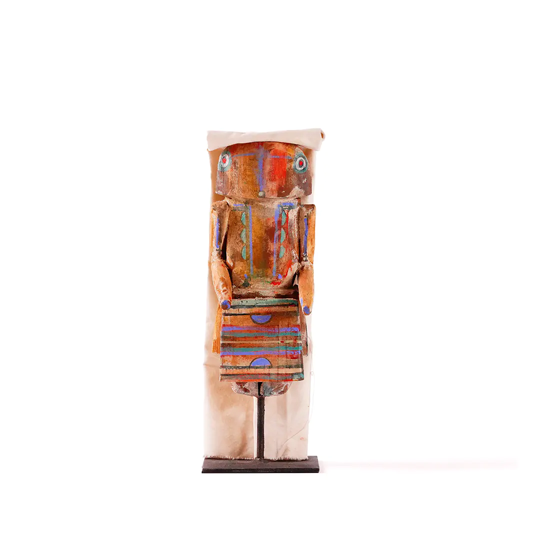 Short, stylized wooden figure with wide painted eyes, weathered orange and blue details, and a striped geometric panel at the front, mounted on a black stand and partially wrapped in pale fabric against a white background.