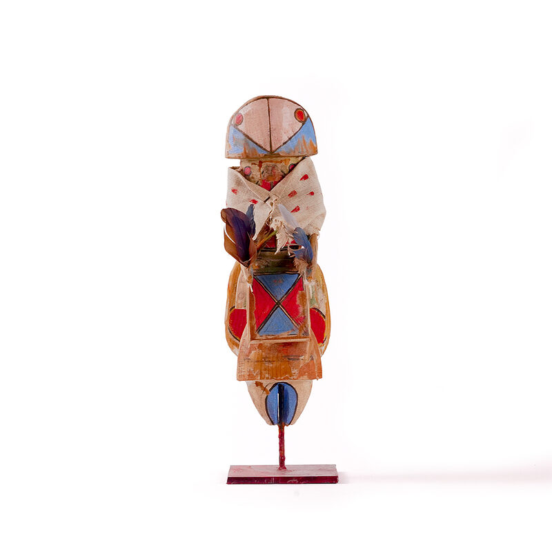 Small stylized Yei sculpture by Sheldon Harvey with painted geometric details, cloth wrap, feathers, and metal base.