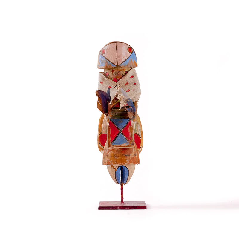 Small stylized Yei sculpture by Sheldon Harvey with painted geometric details, cloth wrap, feathers, and metal base.