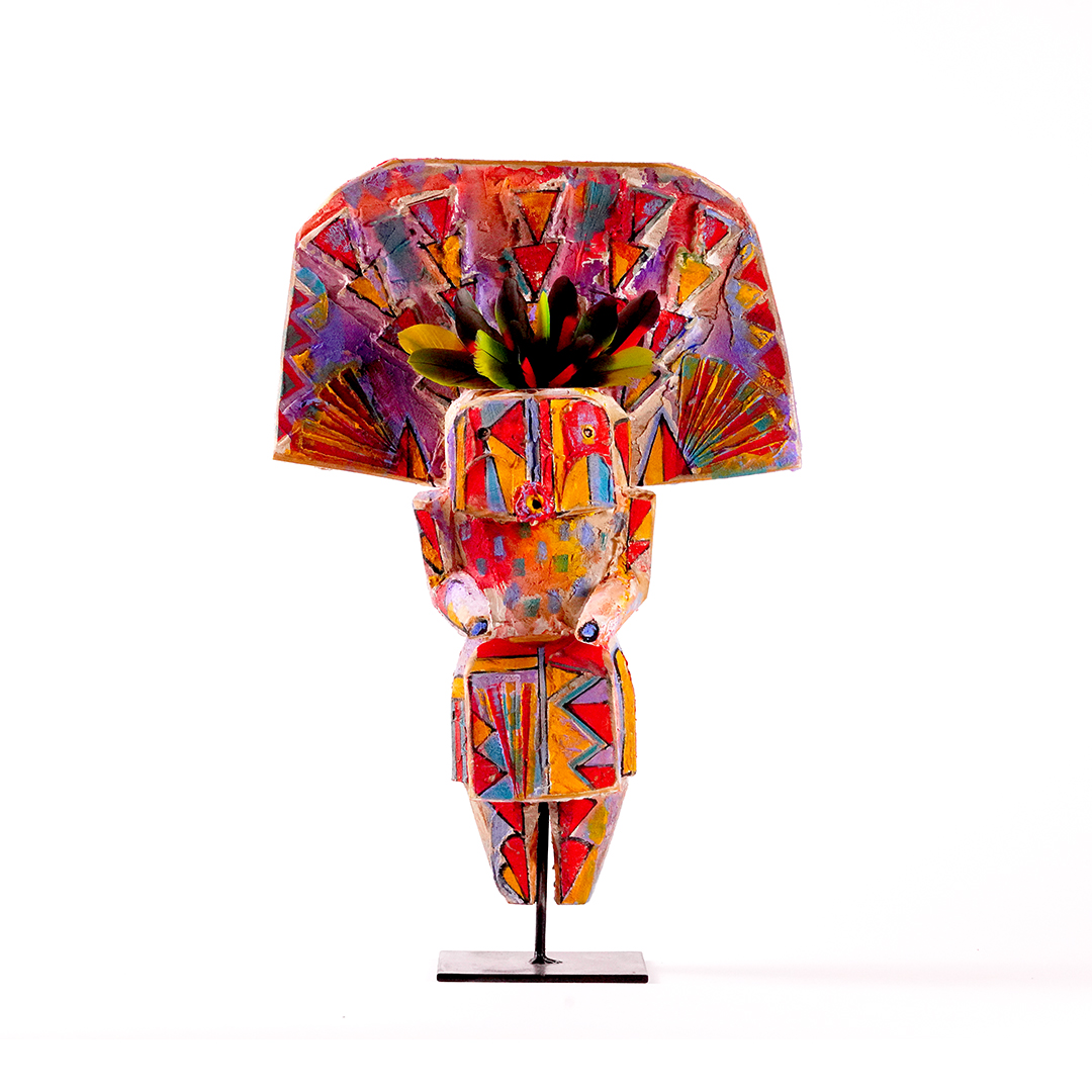 olorful carved wood sculpture titled Yei by Sheldon Harvey, showing a stylized human figure with a wide fan-shaped headdress, geometric painted patterns, and a black floral or feather-like form at the top, mounted on a metal stand against a white background.